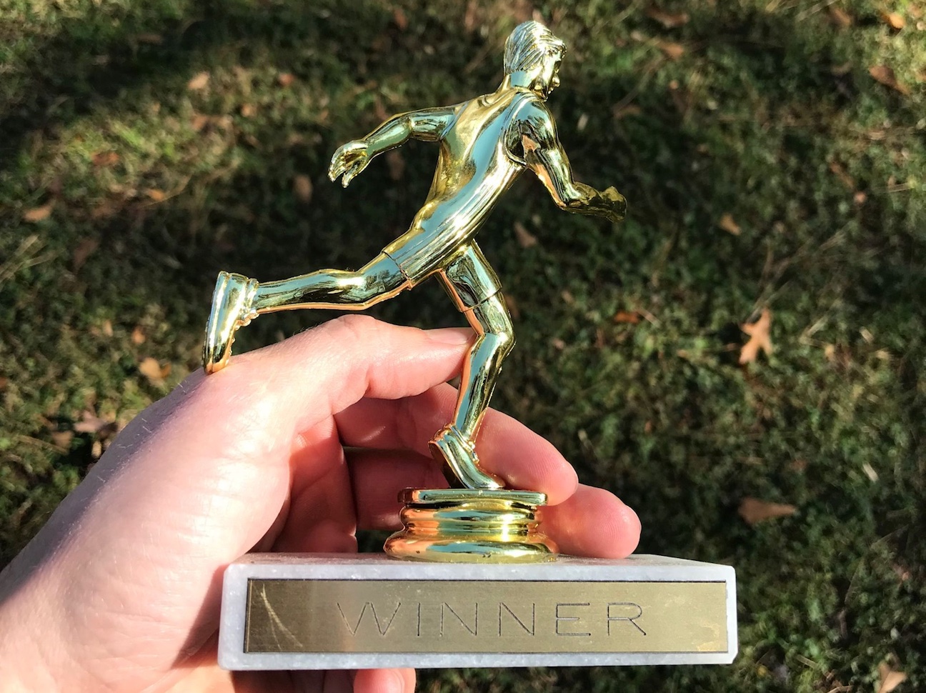 A gold-colored plastic trophy of a runner is held in someone's left hand. The label on the statue says 'WINNER'.