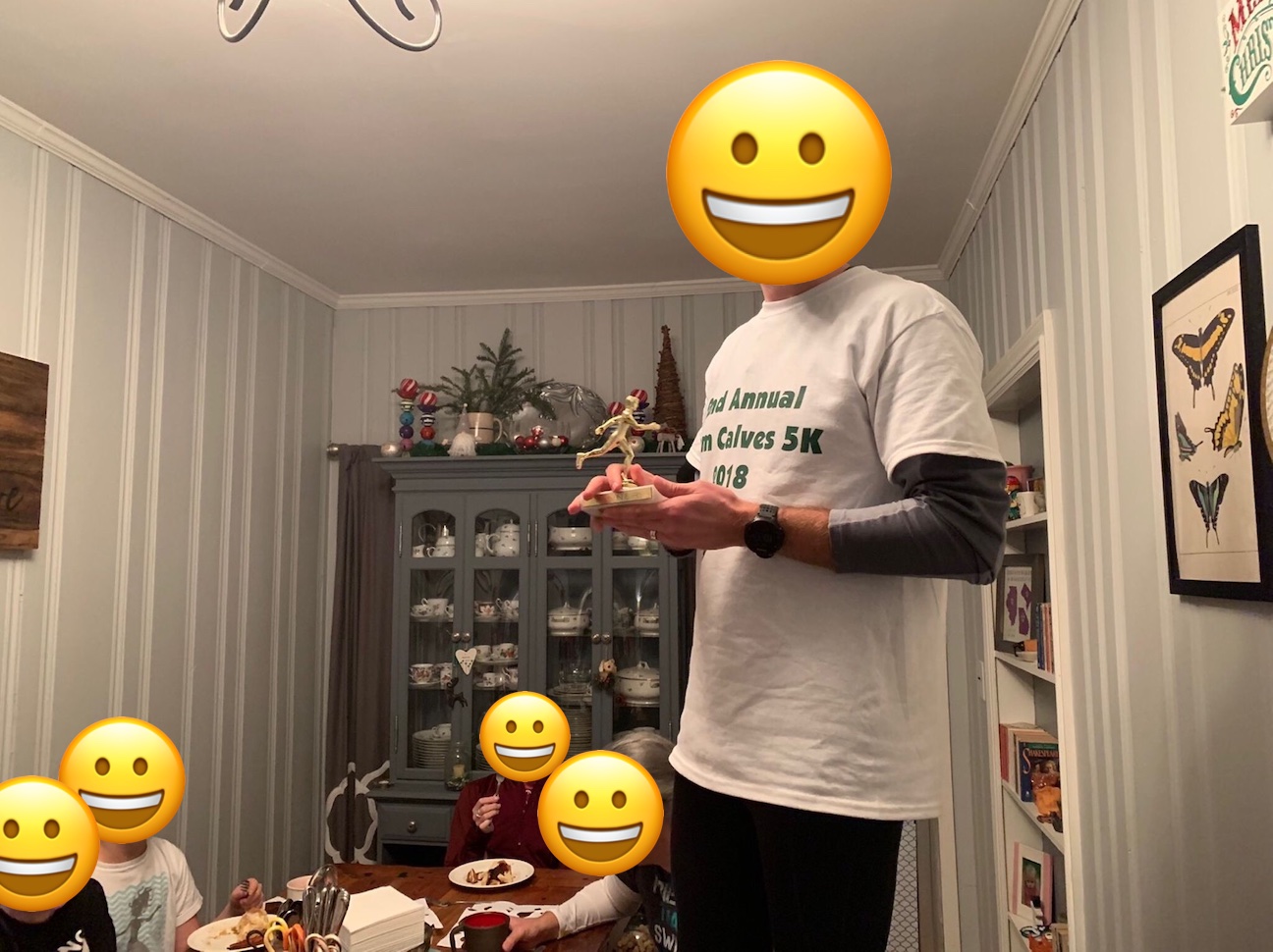 People are eating at a dining room table. One person is standing on a chair, holding a trophy and giving a speech.