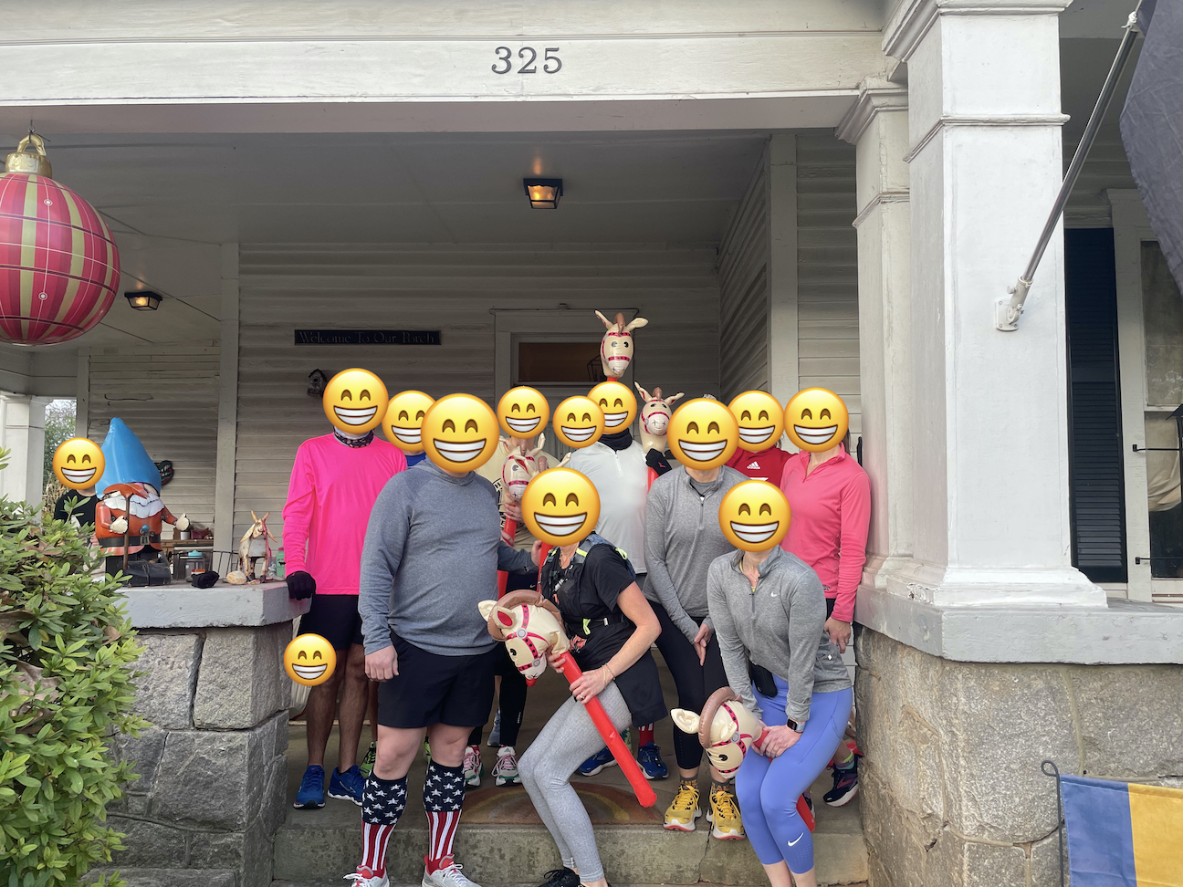 A group of people in running cloths standing on the front porch of a house. Some are holding inflatable hobby horses.