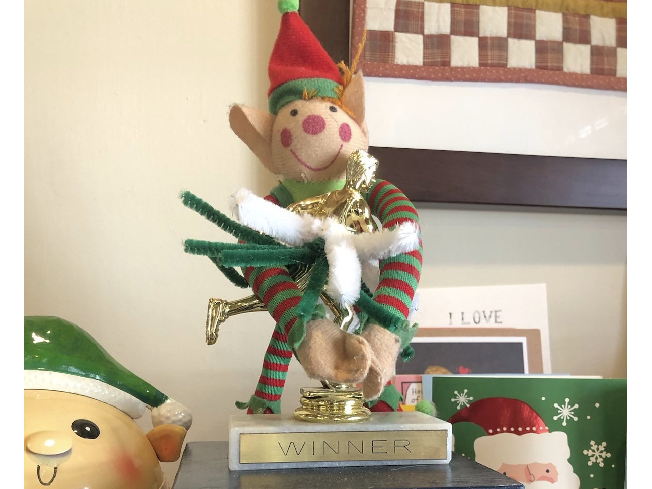 A trophy of a runner with a pipe cleaner skirt and the word “WINNER” being hugged by a stuffed elf.